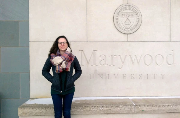 Woman in a scarf and coat standing in front of a Marywood University sign on a stone wall.
